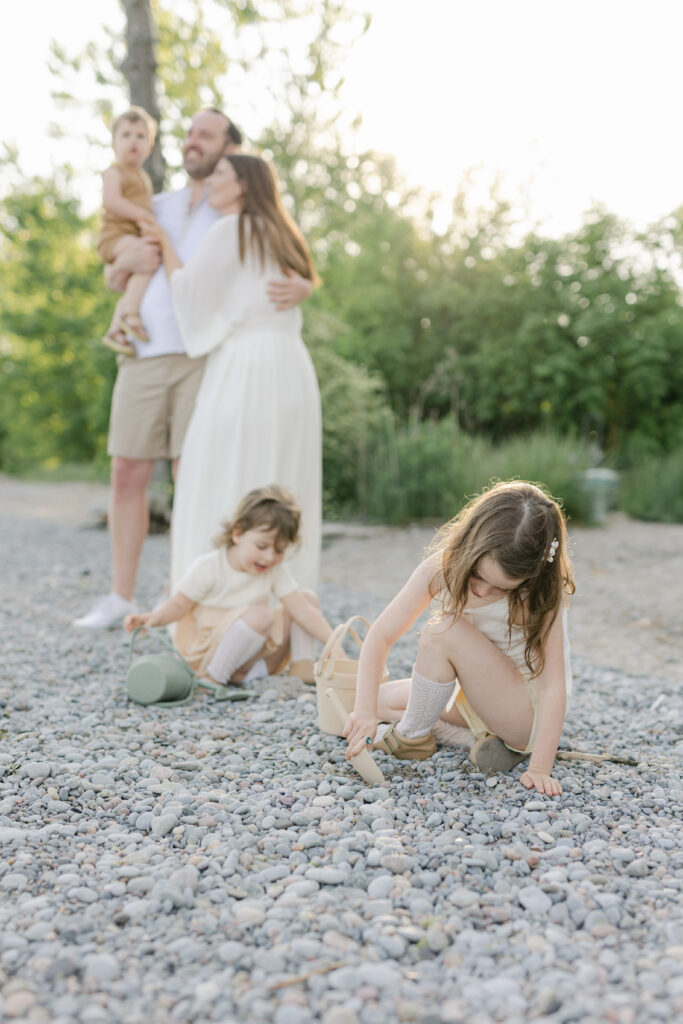 Two toddler sisters play in the rocks on the beach during their Toronto Family Session with mom and dad behind them 