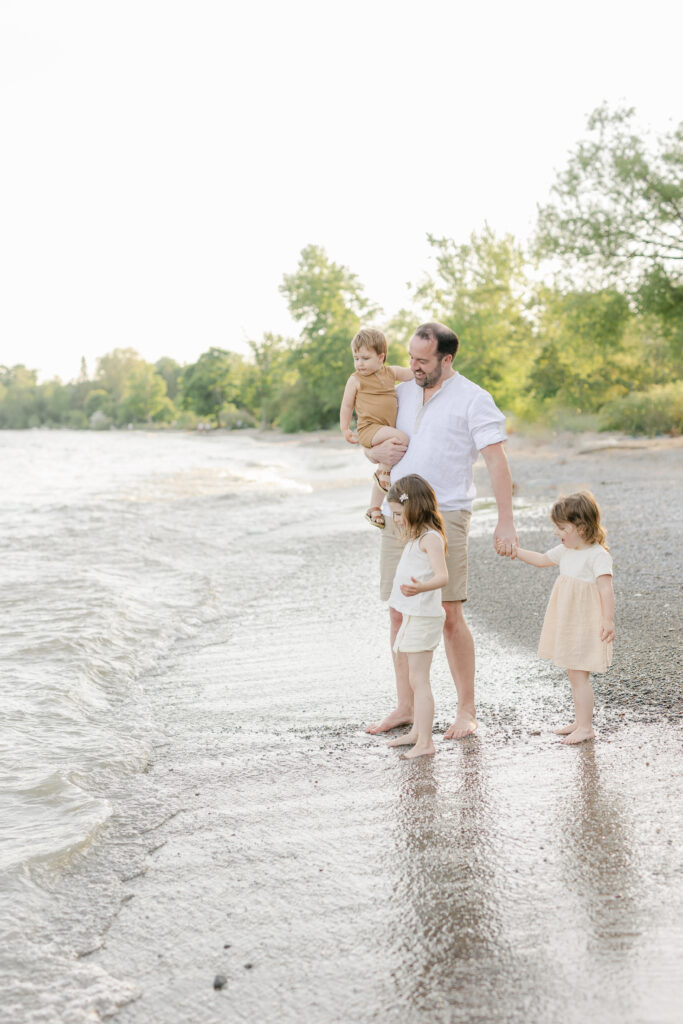 A father explores the water on the beach with his two toddler daughters and younger son during a Toronto Family Session