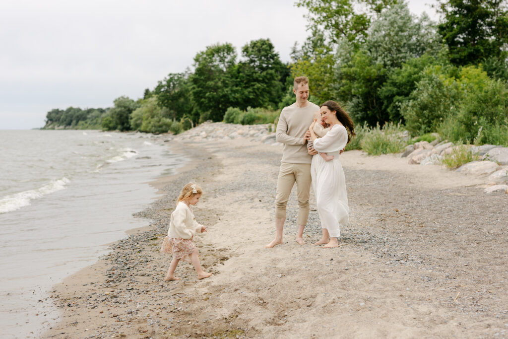 A little girl walks on the beach while mom and dad are hugging and watching her