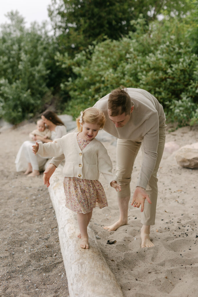 A dad is helping his daughter walk on a tree log while mom hugs her baby in the background