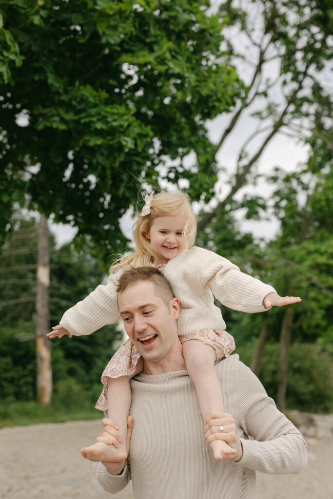 A dad is carrying his daughter on his shoulders and smiling at a Toronto beach